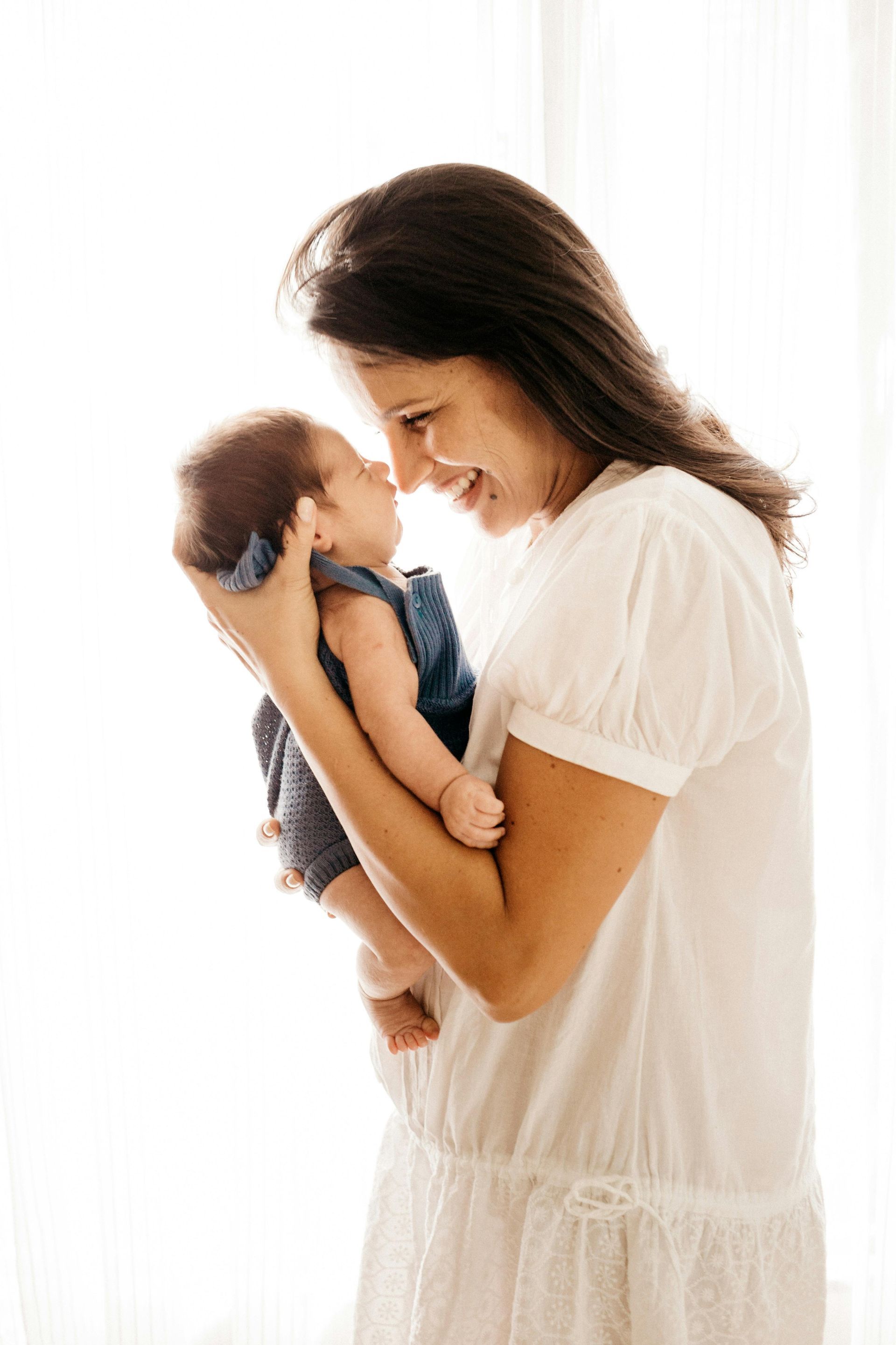 Woman is holding a small baby to her face. She is smiling and standing in front of a white curtain.