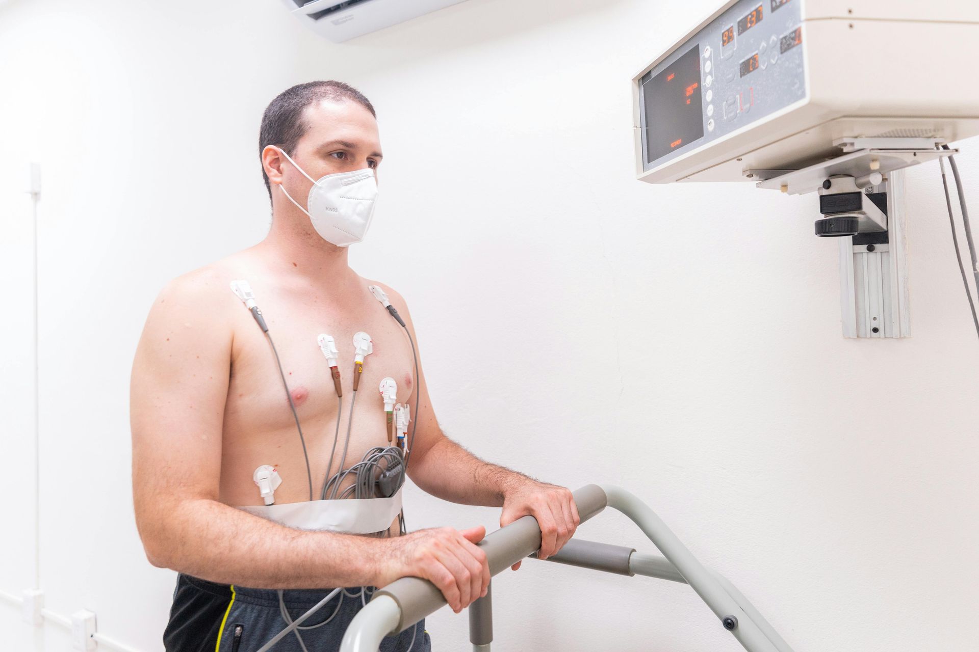 Man with chest sensors on a treadmill, wearing a mask, near a monitoring device.