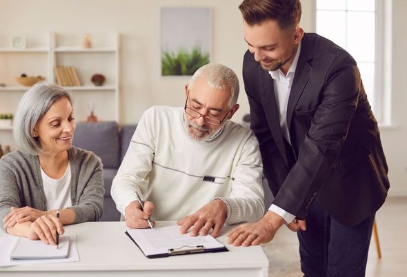 An advisor stands beside an individual who is signing a document at a desk while another person watches.