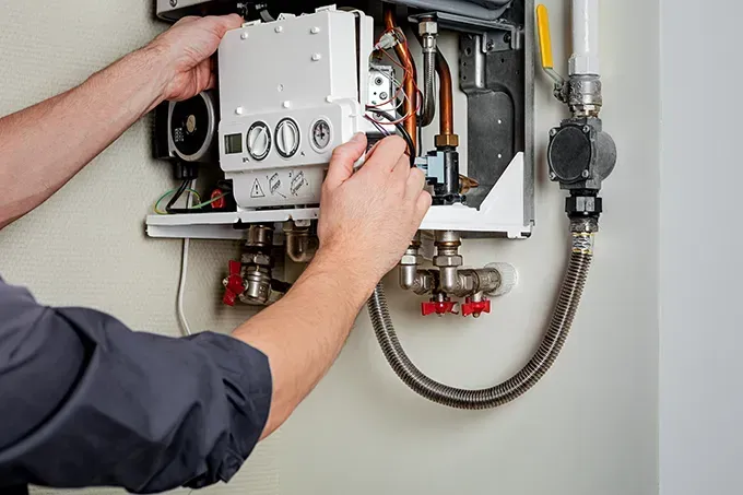 Technician repairing an open wall-mounted boiler with exposed pipes and controls