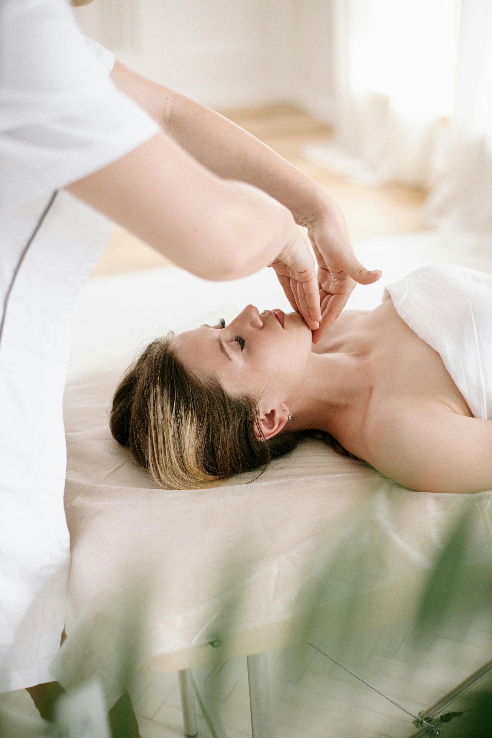 Person receiving a facial massage in a bright room. Hands on jawline, white linens, and relaxed expression.