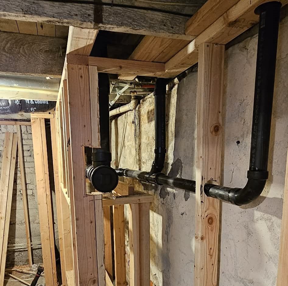 Construction site showing exposed black PVC plumbing pipes inside wall studs and a trench cut into a concrete floor.