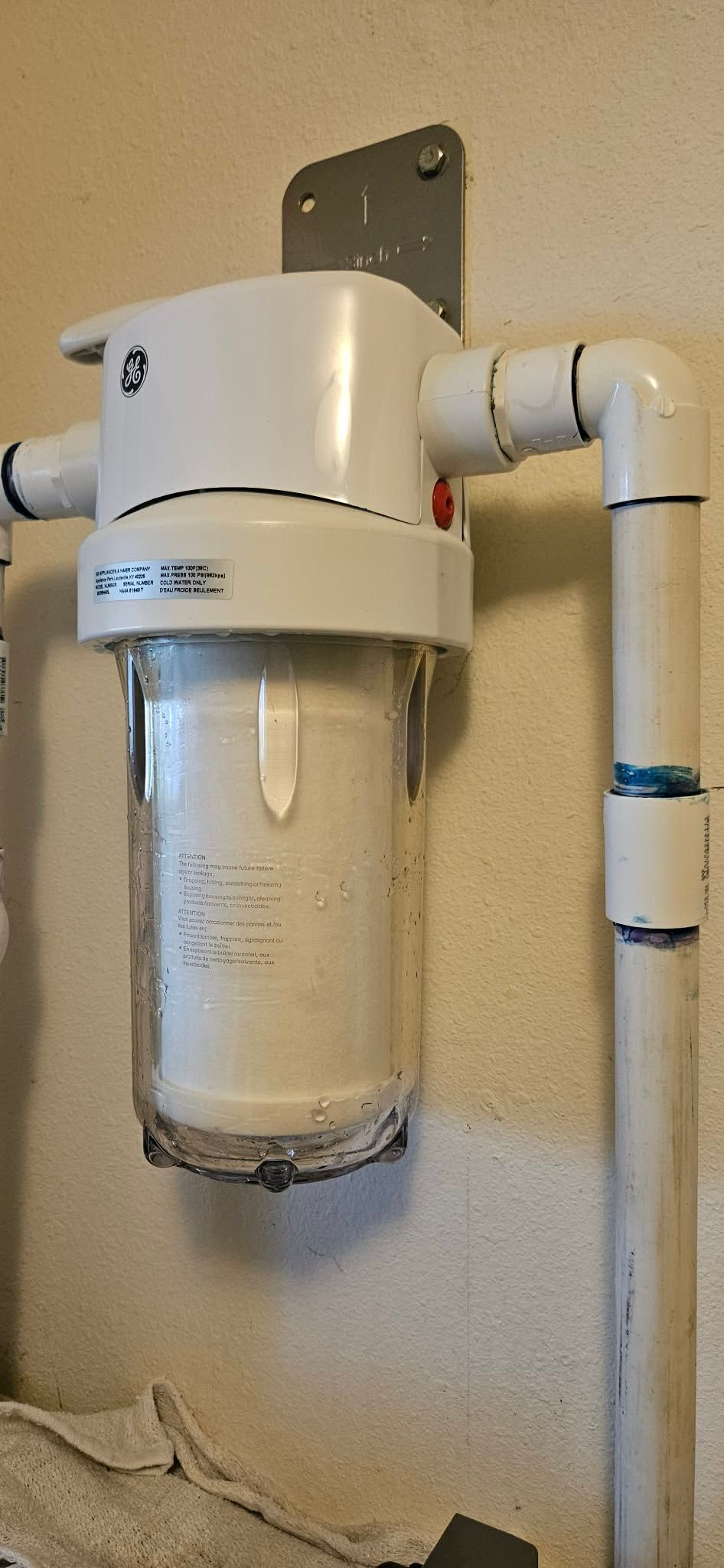 A GE brand water filter system mounted on a wall with white PVC plumbing pipes connected to the sides.