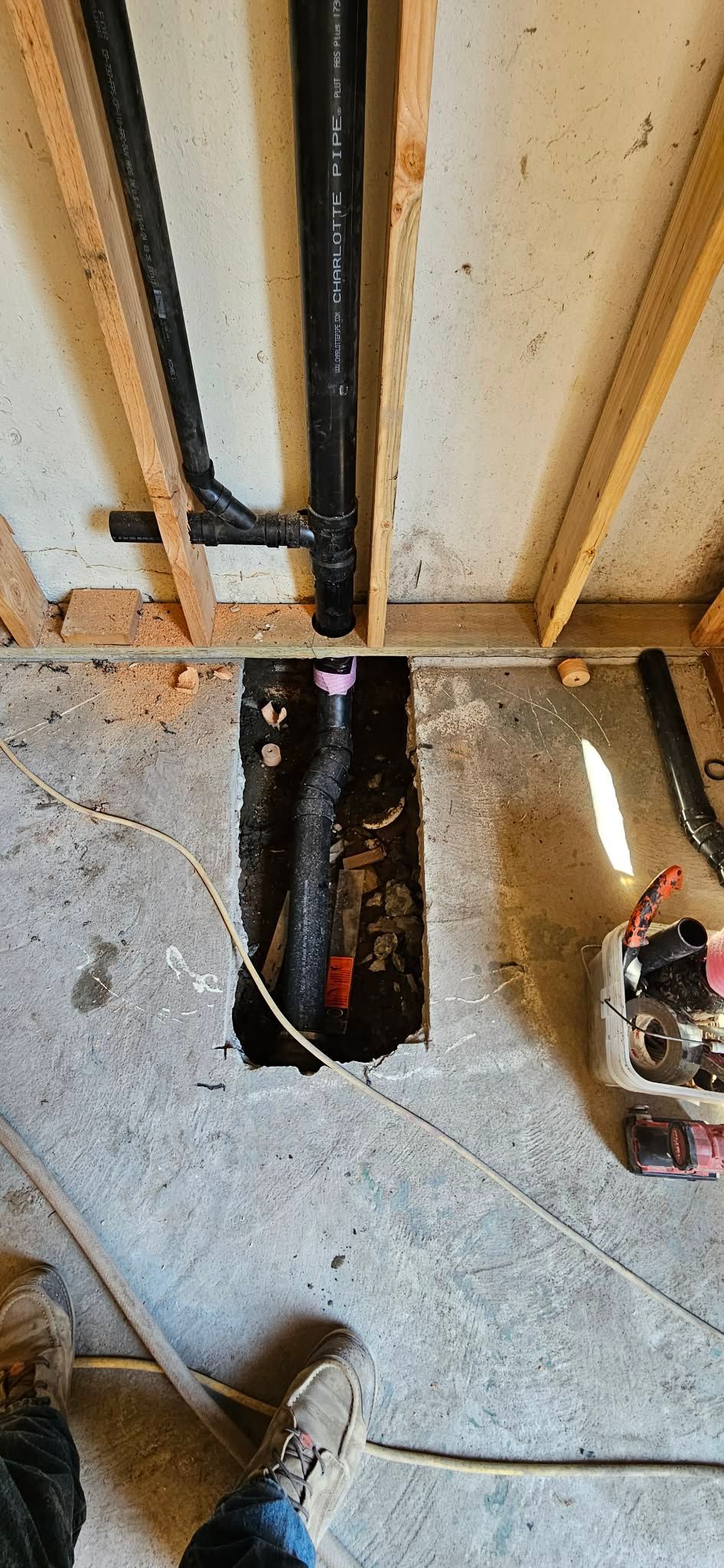 Construction site showing exposed black PVC plumbing pipes inside wall studs and a trench cut into a concrete floor.