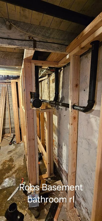 Exposed plumbing pipes installed in the wooden wall framing of a basement bathroom under construction.