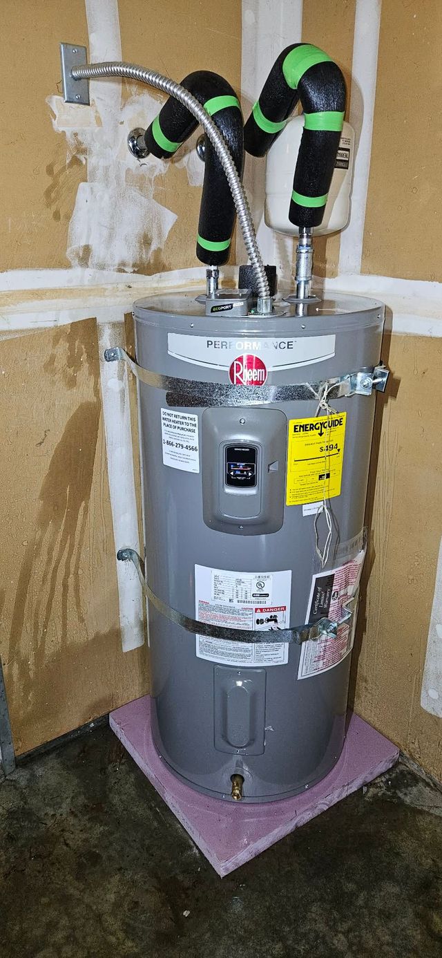 A gray Rheem electric water heater sits in a corner on a pink insulation board with insulated pipes connected to the top.