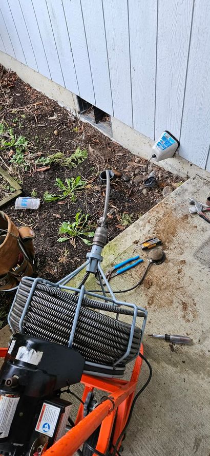 An orange plumbing auger, used for drain cleaning, sits on a concrete patio near a house exterior wall.
