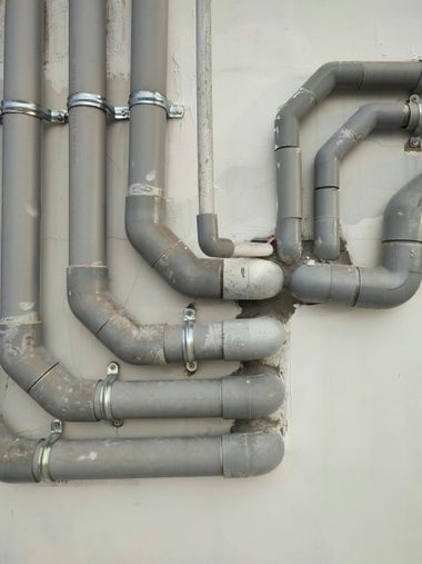Multiple grey conduit pipes mounted against a plain wall, featuring several sharp 90-degree elbows and metal support clamps.