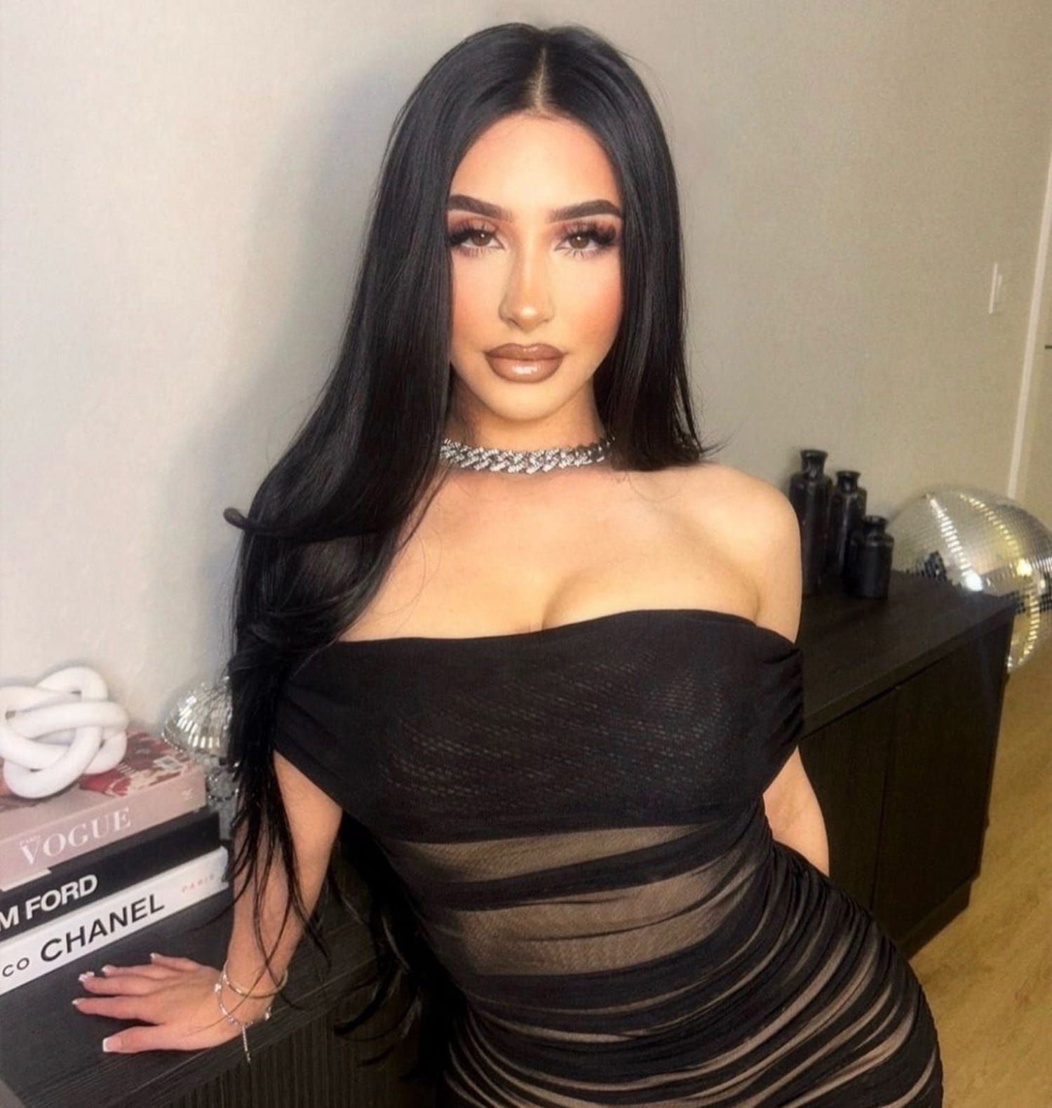 Woman in a black off-shoulder dress posing indoors beside a dresser and stacked books.