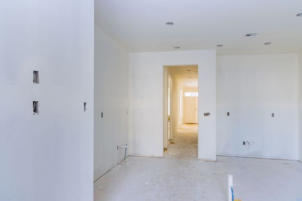 Interno di una stanza in costruzione, pareti dipinte di bianco, con prese elettriche e una porta che conduce a un corridoio.