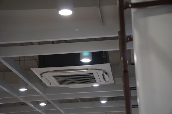 Unità di aria condizionata montata a soffitto. Scatola nera e griglia bianca. Interno con travi, luci e superfici bianco sporco.