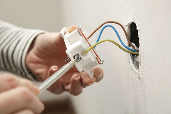 Persona che installa una presa elettrica usando un cacciavite. I fili sono marroni, blu e giallo-verdi.