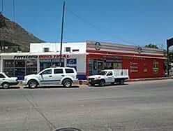 Una ferretería con exterior rojo y blanco, vehículos estacionados y una montaña al fondo.