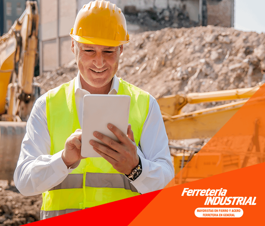 Trabajador de la construcción con chaleco amarillo y casco usando una tableta en el sitio; excavadora y tierra en el fondo.