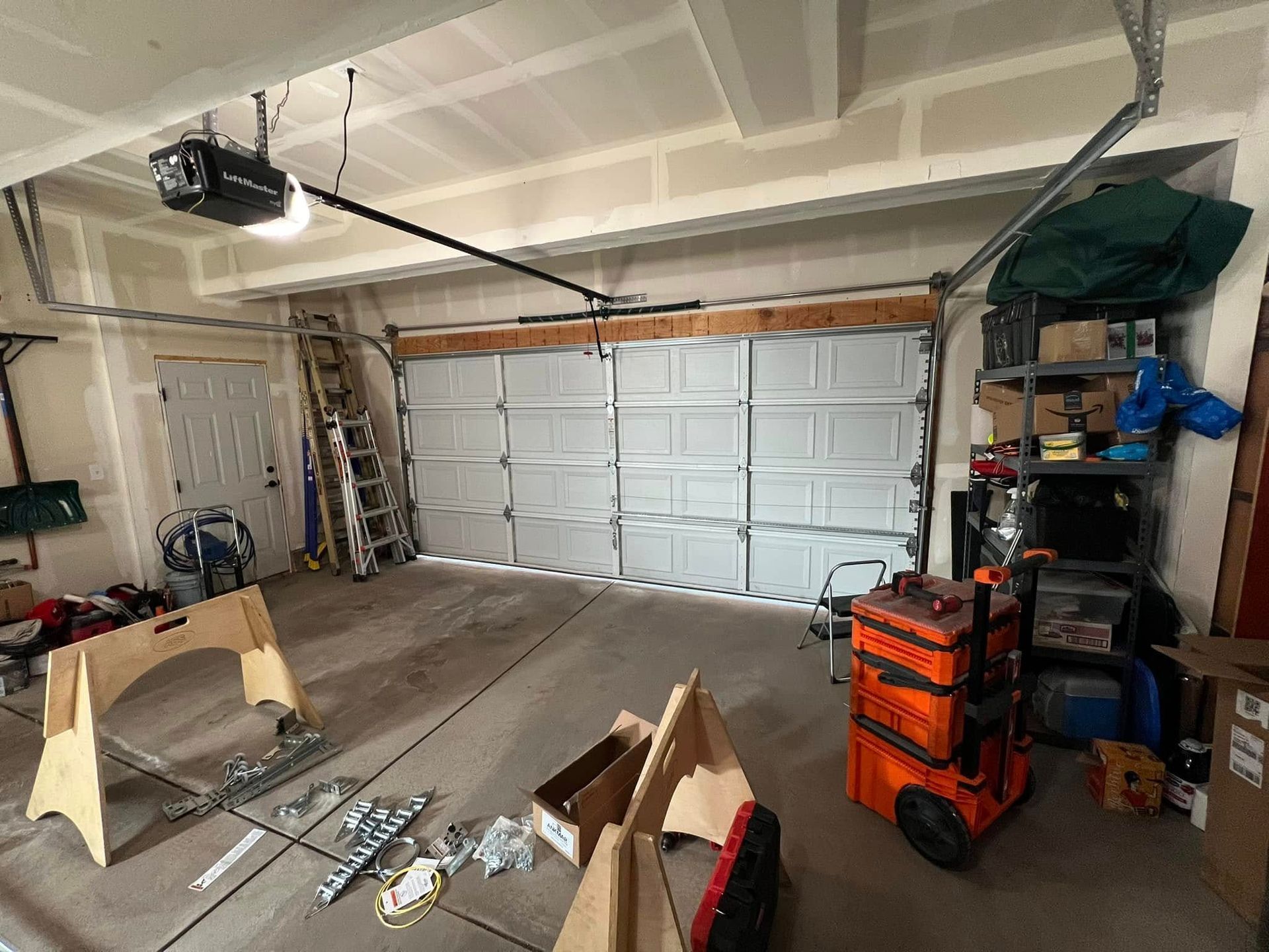 A garage interior featuring a white sectional door, a rolling orange toolbox, sawhorses, and miscellaneous tools.