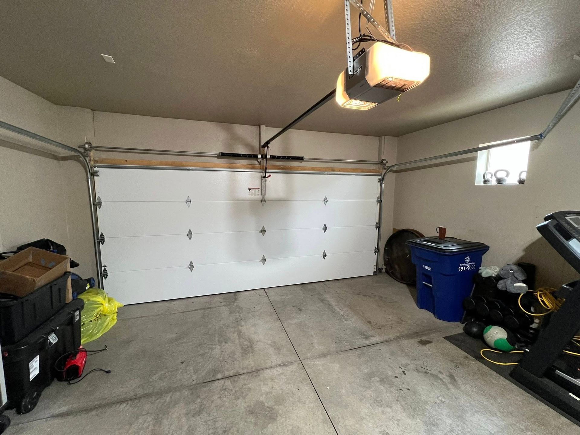 A white garage door is closed inside a beige garage, with a ceiling opener, concrete floor, blue bin, and gym equipment.