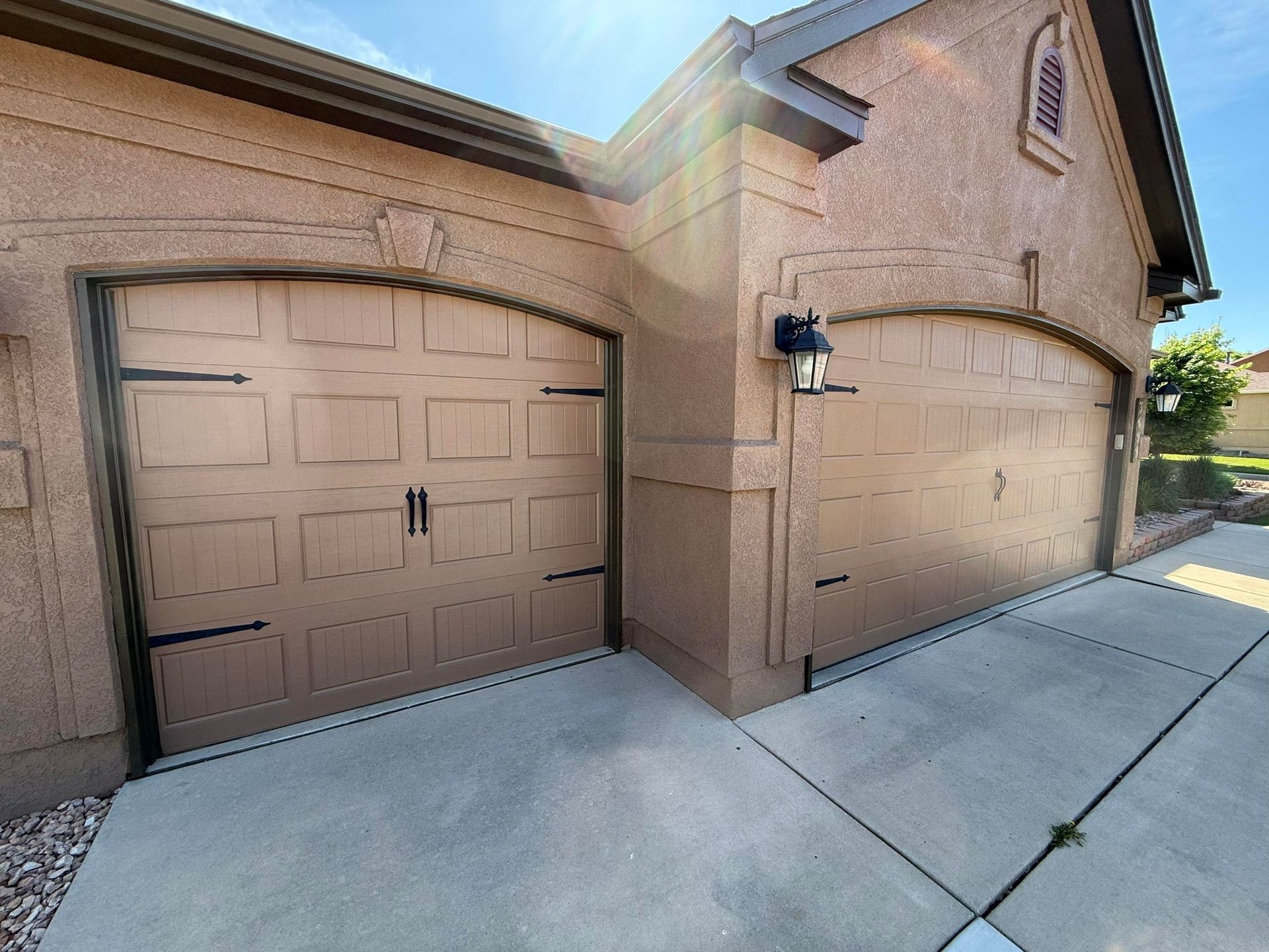A two-car garage with arched, tan paneled doors, black decorative hinges, and stucco walls under a bright blue sky.