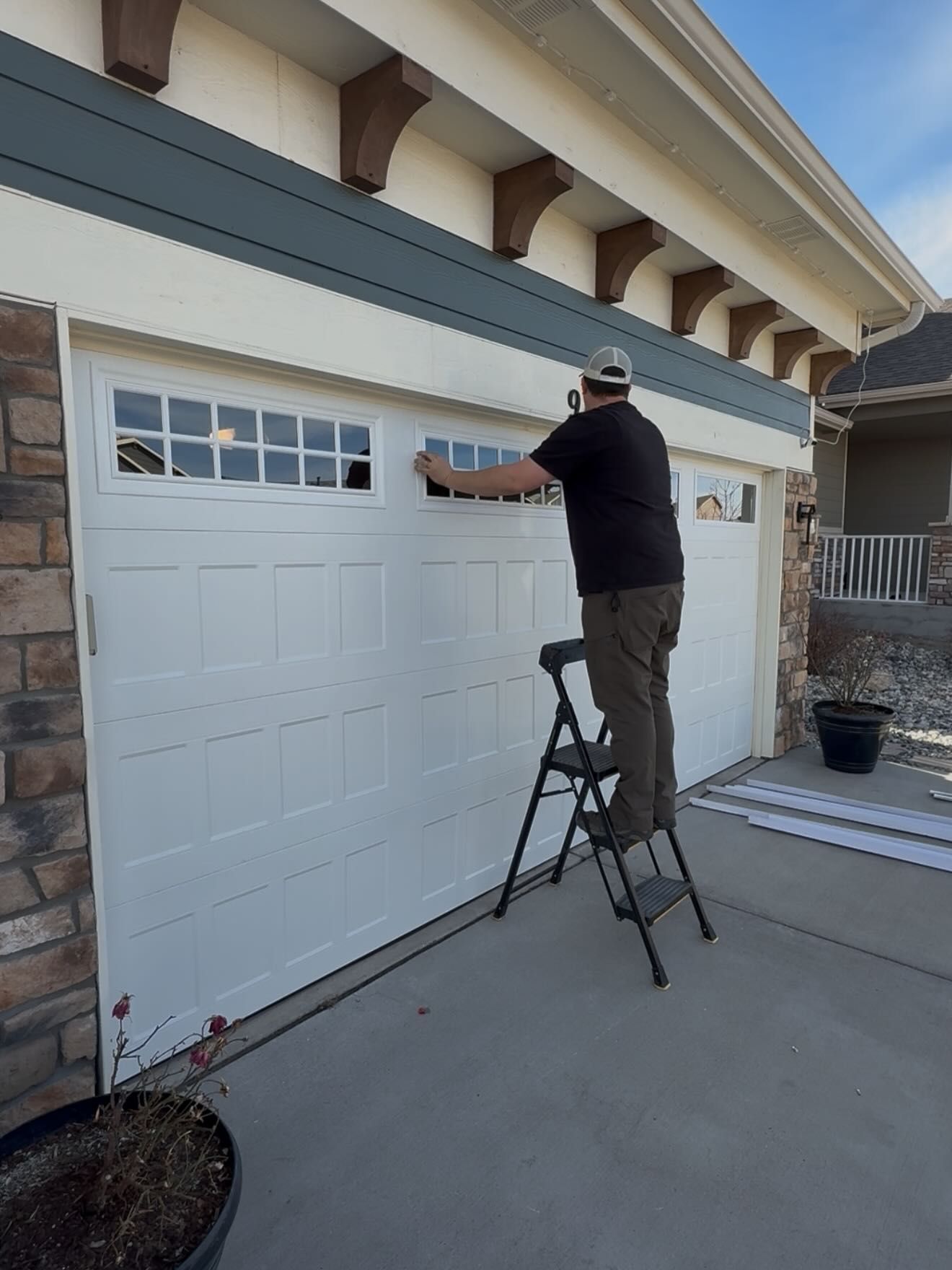 A person standing on a step ladder, installing or adjusting decorative window inserts onto a white garage door.