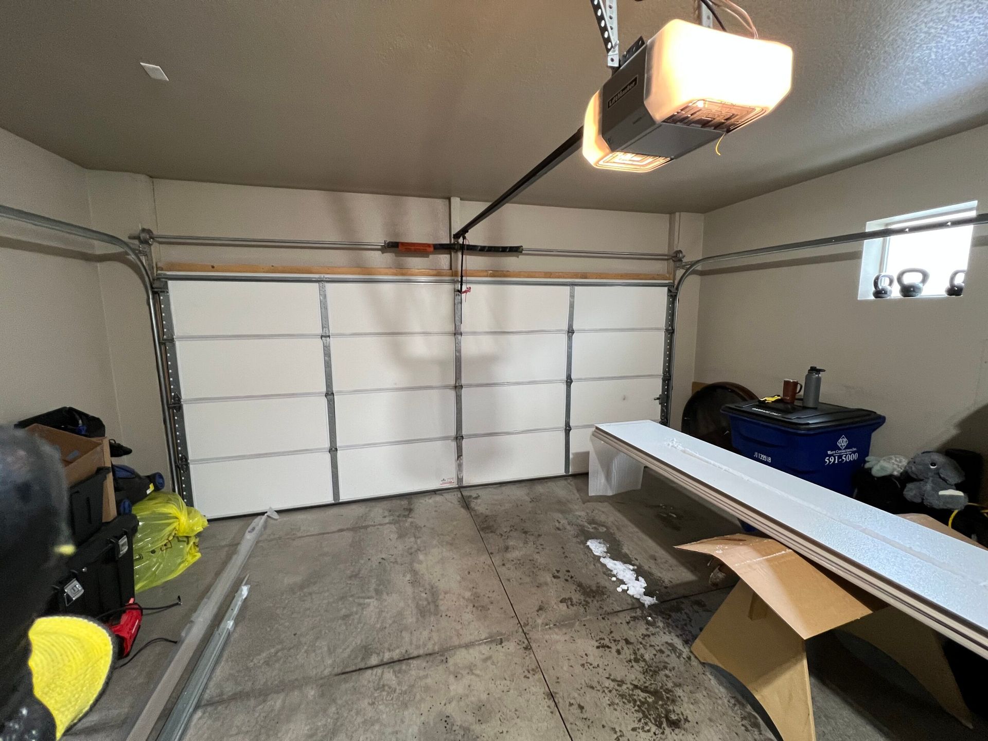 The inside of a garage featuring a white overhead door, a ceiling-mounted opener, and various stored items.