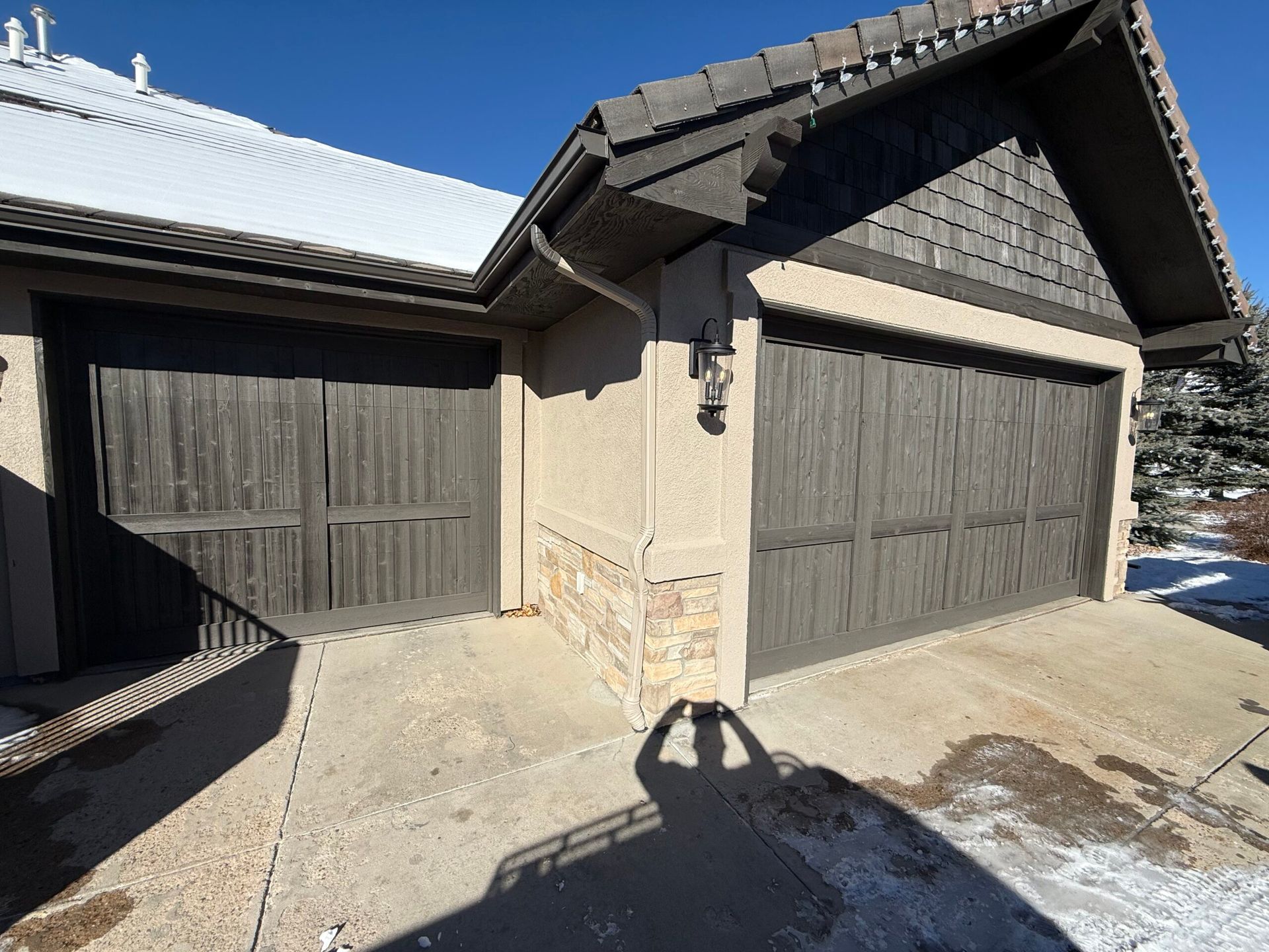 A house with two dark wooden garage doors, beige stucco walls, stone accents, and a brown shingled roof on a sunny day.