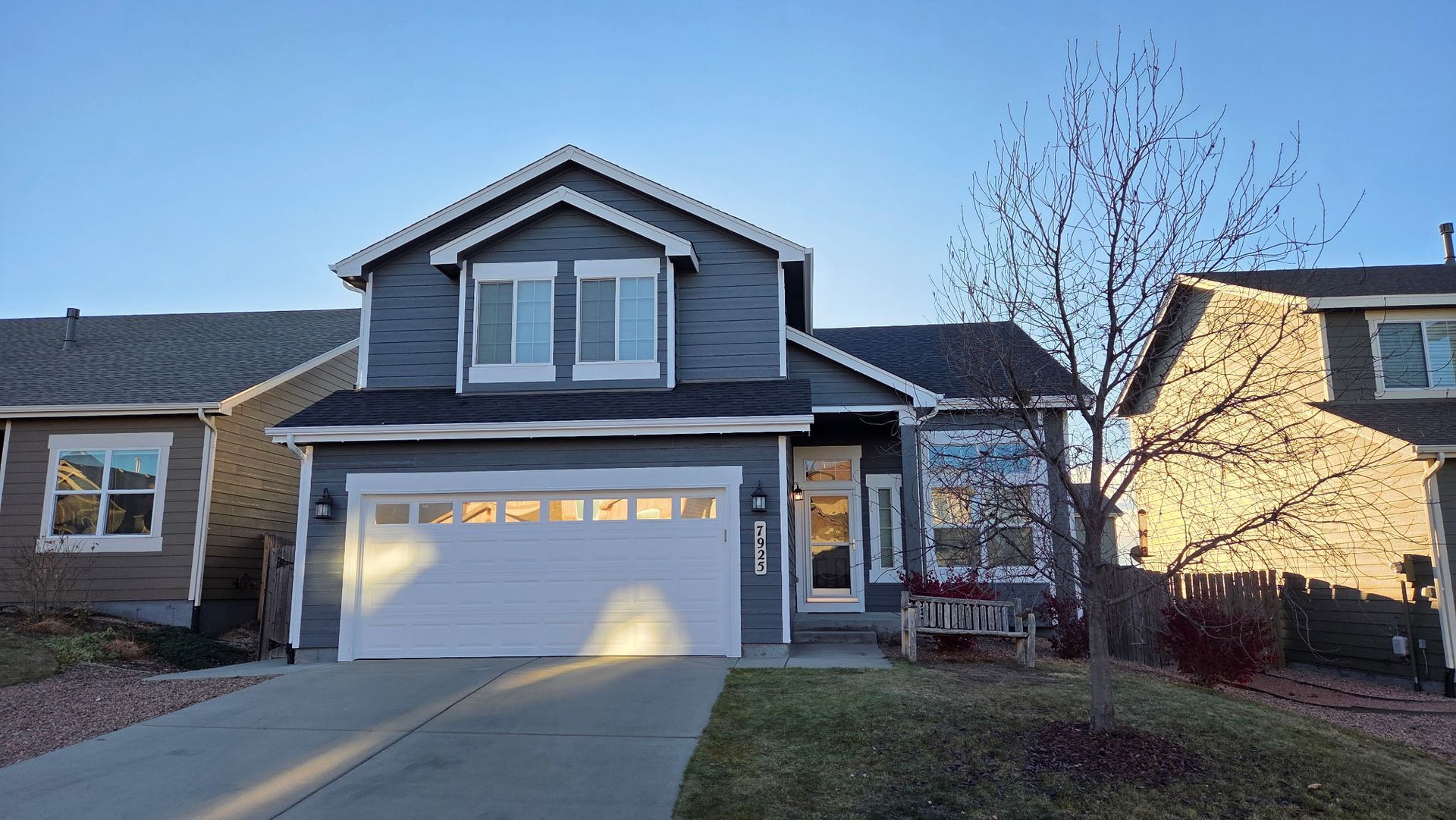 A two-story grey residential house with a white garage door, concrete driveway, and a bare tree in the front yard.