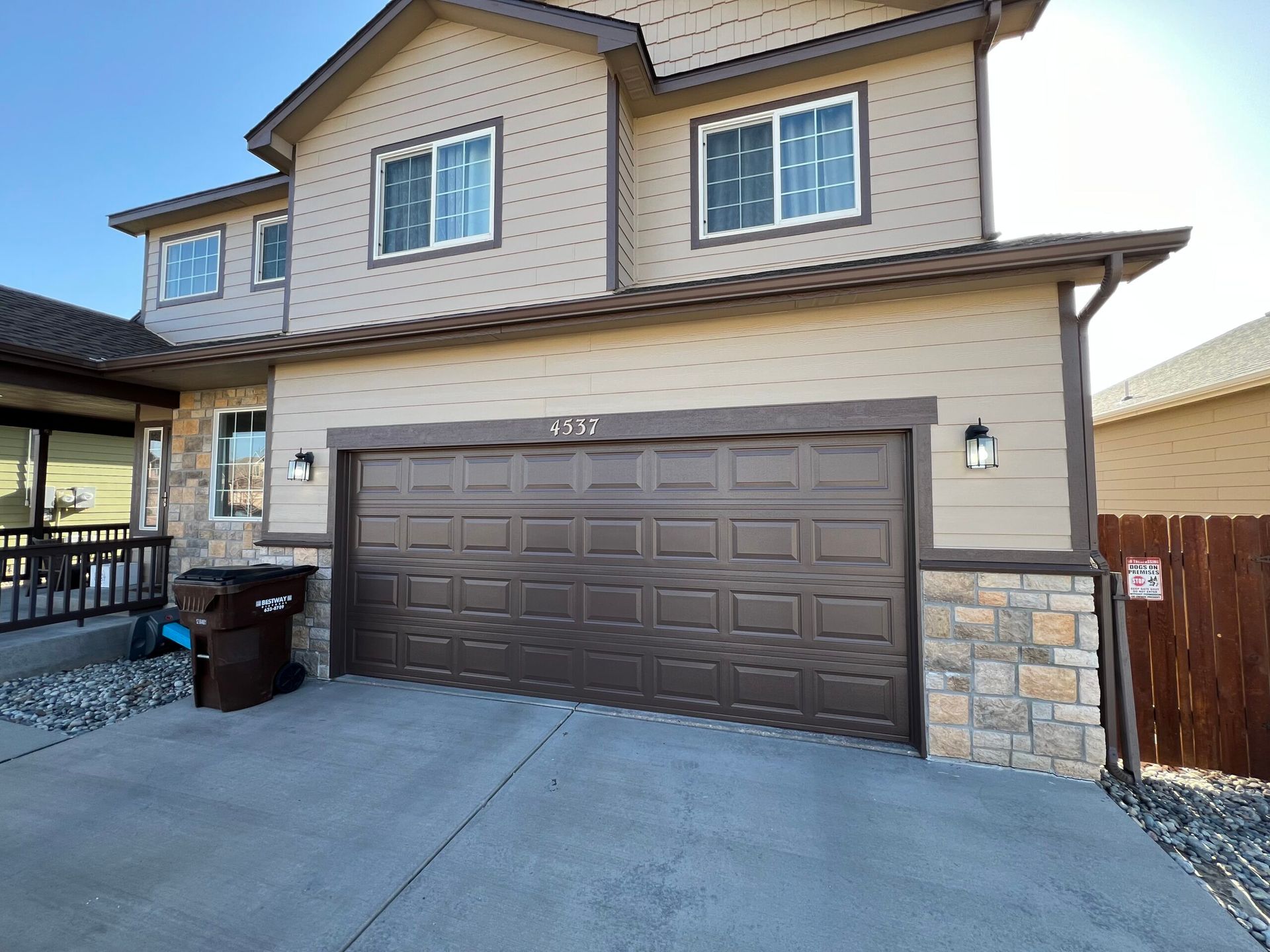 A two-story tan house with a dark brown garage door, stone accents on the lower facade, and a small porch on the left.