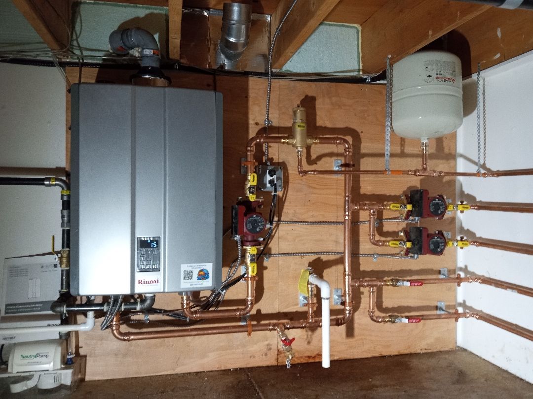A wall-mounted silver tankless water heater with copper pipes, pumps, and a white expansion tank in a basement.
