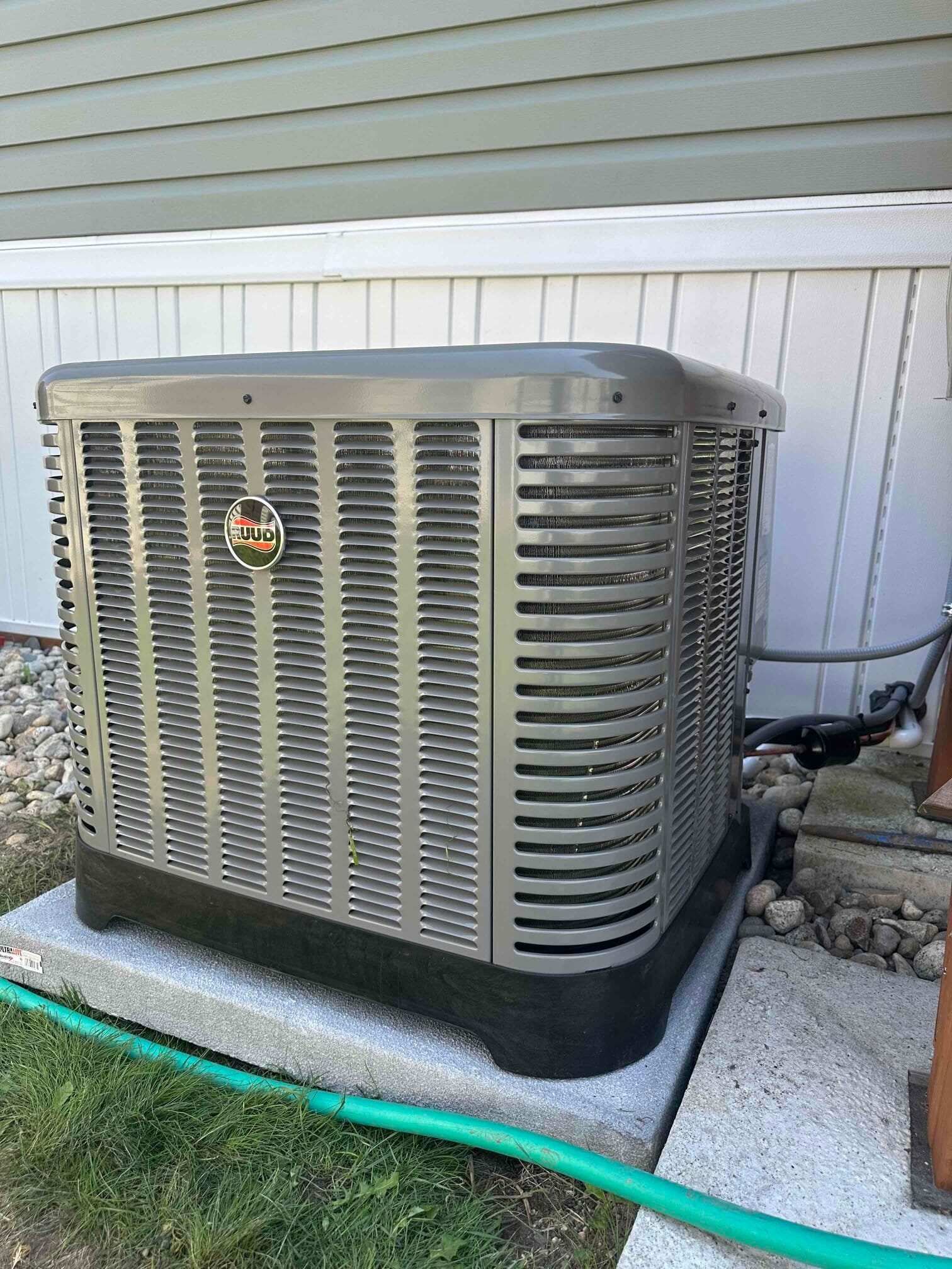 A large air conditioner is sitting outside of a house next to a green hose.