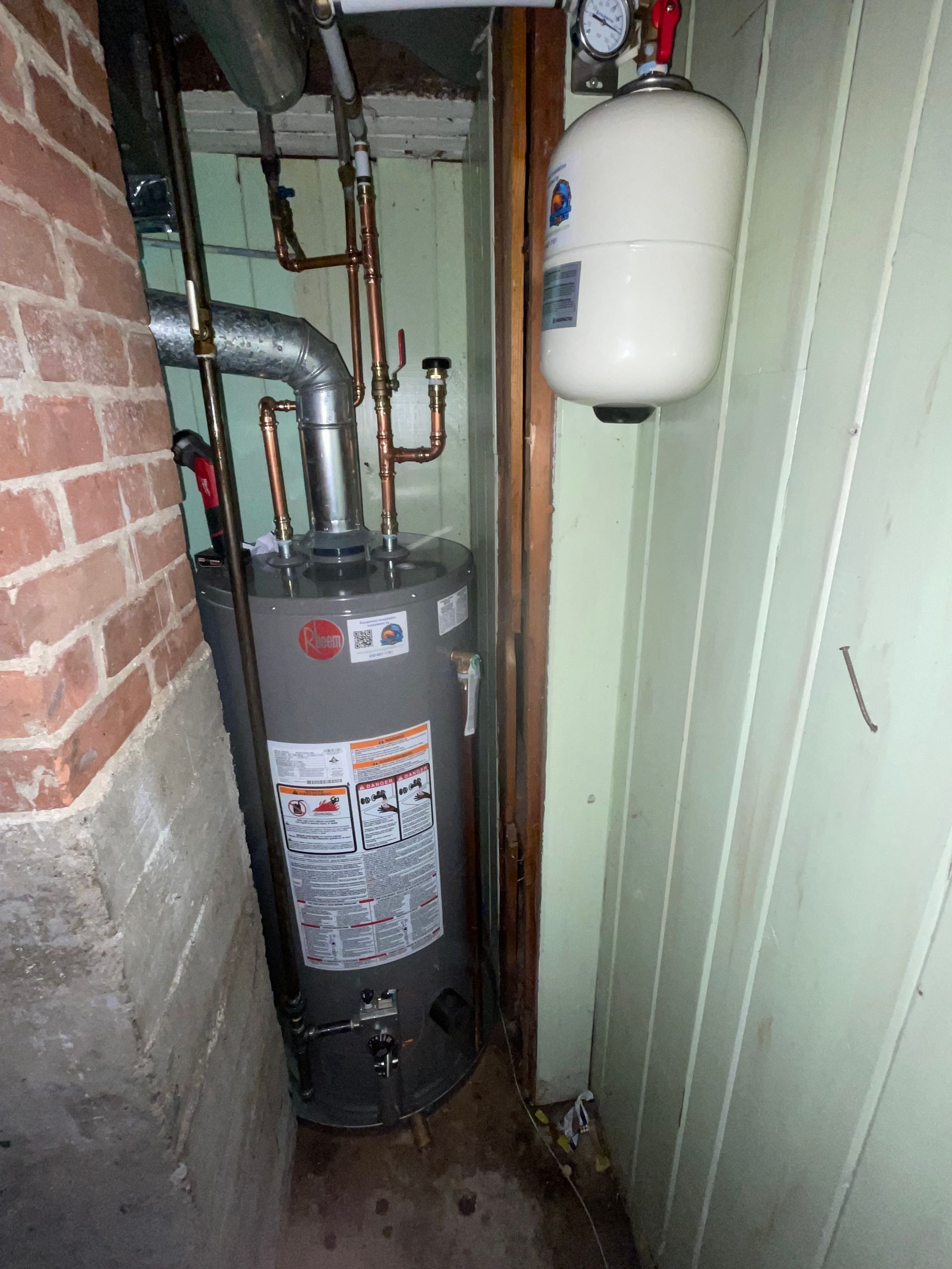 A water heater is sitting in a corner of a room next to a brick wall.