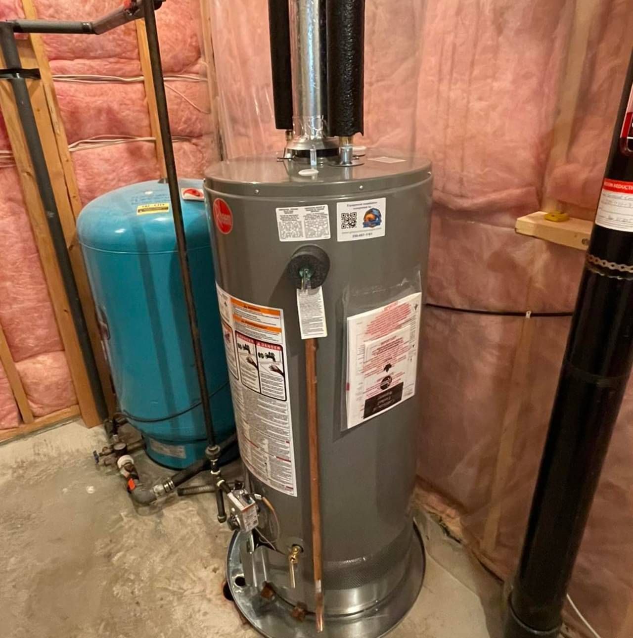 A water heater is sitting in a basement next to a blue tank.