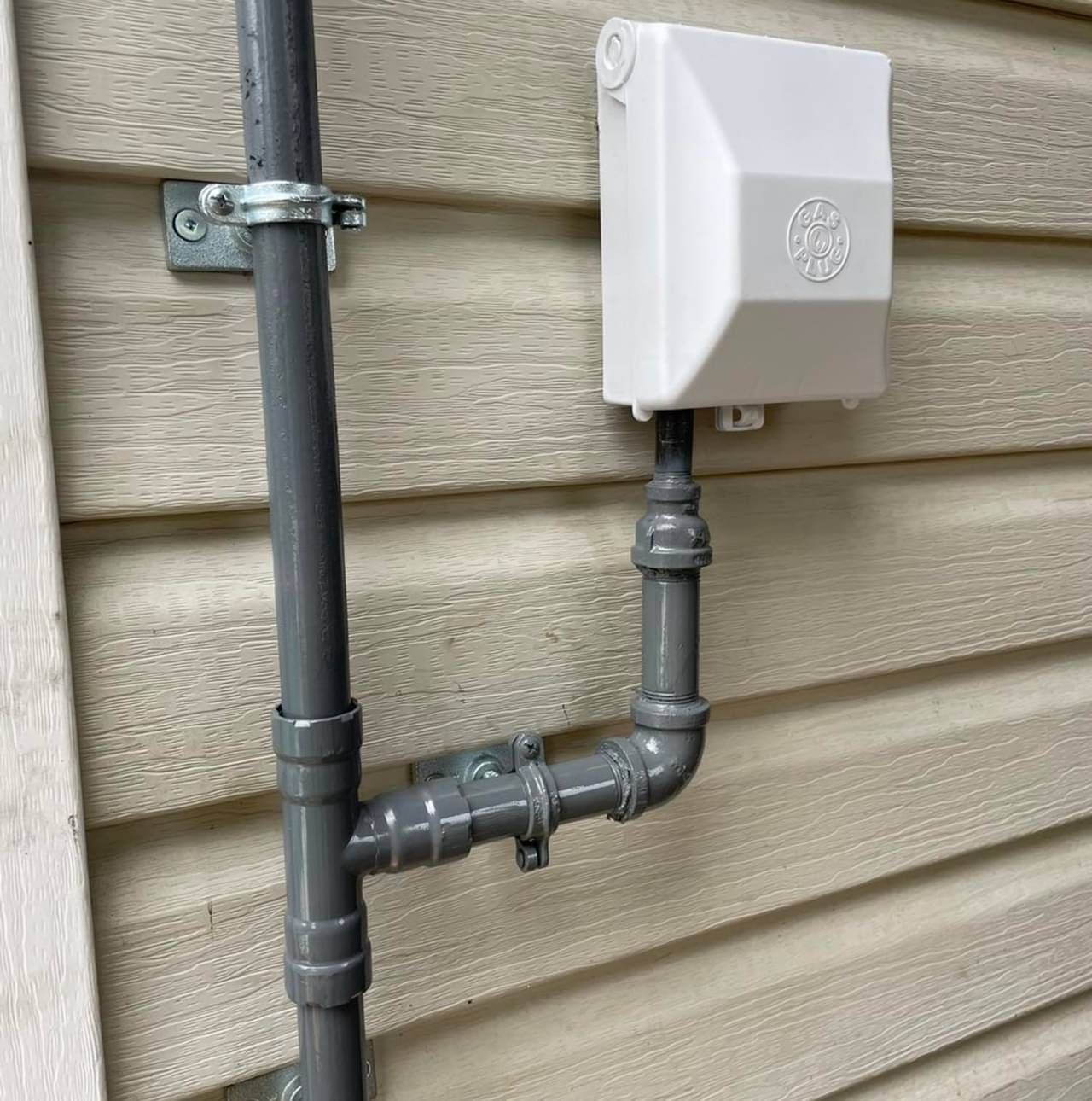 A gray pipe is attached to a white box on the side of a house.