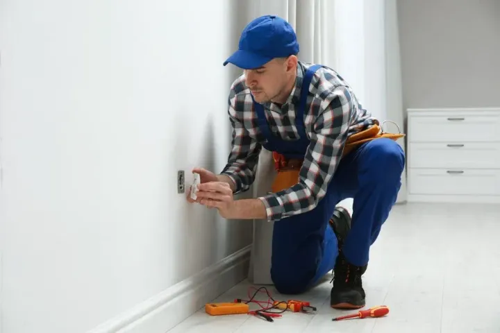 An electrician wearing a plaid shirt and blue overalls kneels to install or repair a wall outlet.