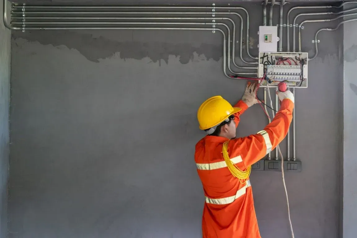 A worker in an orange uniform and yellow hard hat installs electrical wiring into a wall-mounted panel.