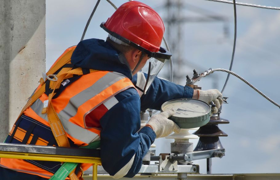 A utility worker in a red hard hat and orange high-visibility vest works on electrical equipment atop a lift.