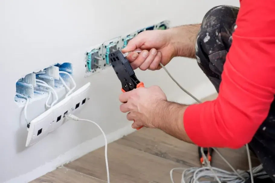 A technician in a red shirt uses a crimping tool to wire Ethernet cables into a wall-mounted network port panel.