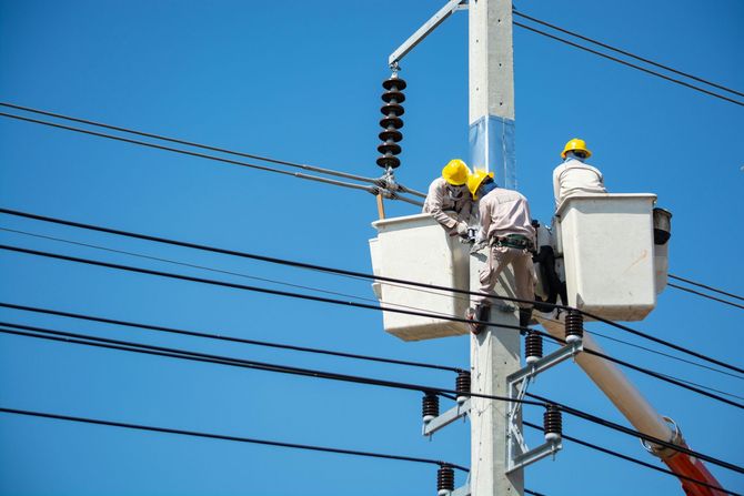Two utility workers in yellow hard hats and white coveralls work on electrical power lines from aerial lift buckets.