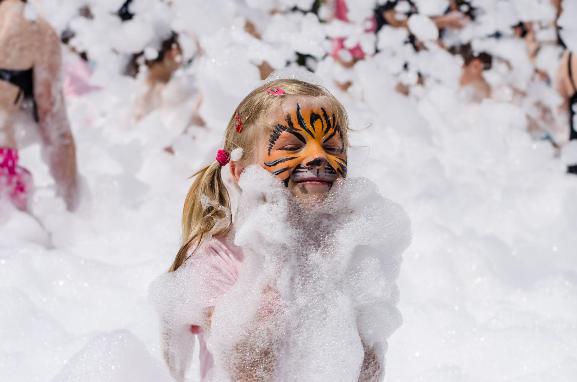 Girl with tiger face paint in a foam party, eyes closed, arms covered in foam.
