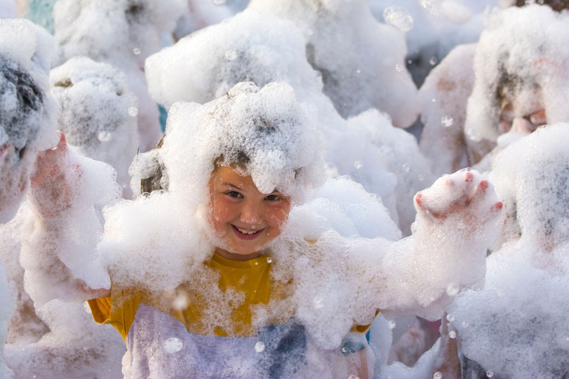 Boy smiling, covered in foam, arms outstretched in fun, at a foam party.