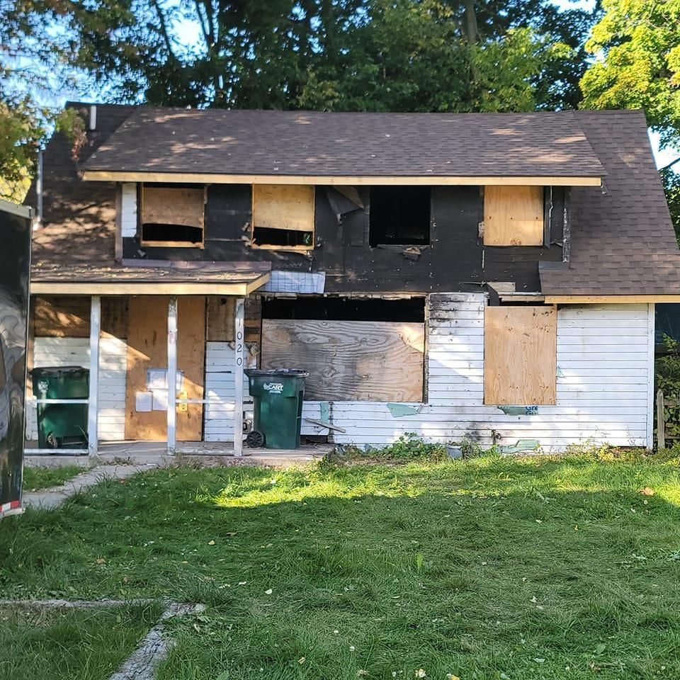 A white house with a brown roof and a green trash can in front of it.