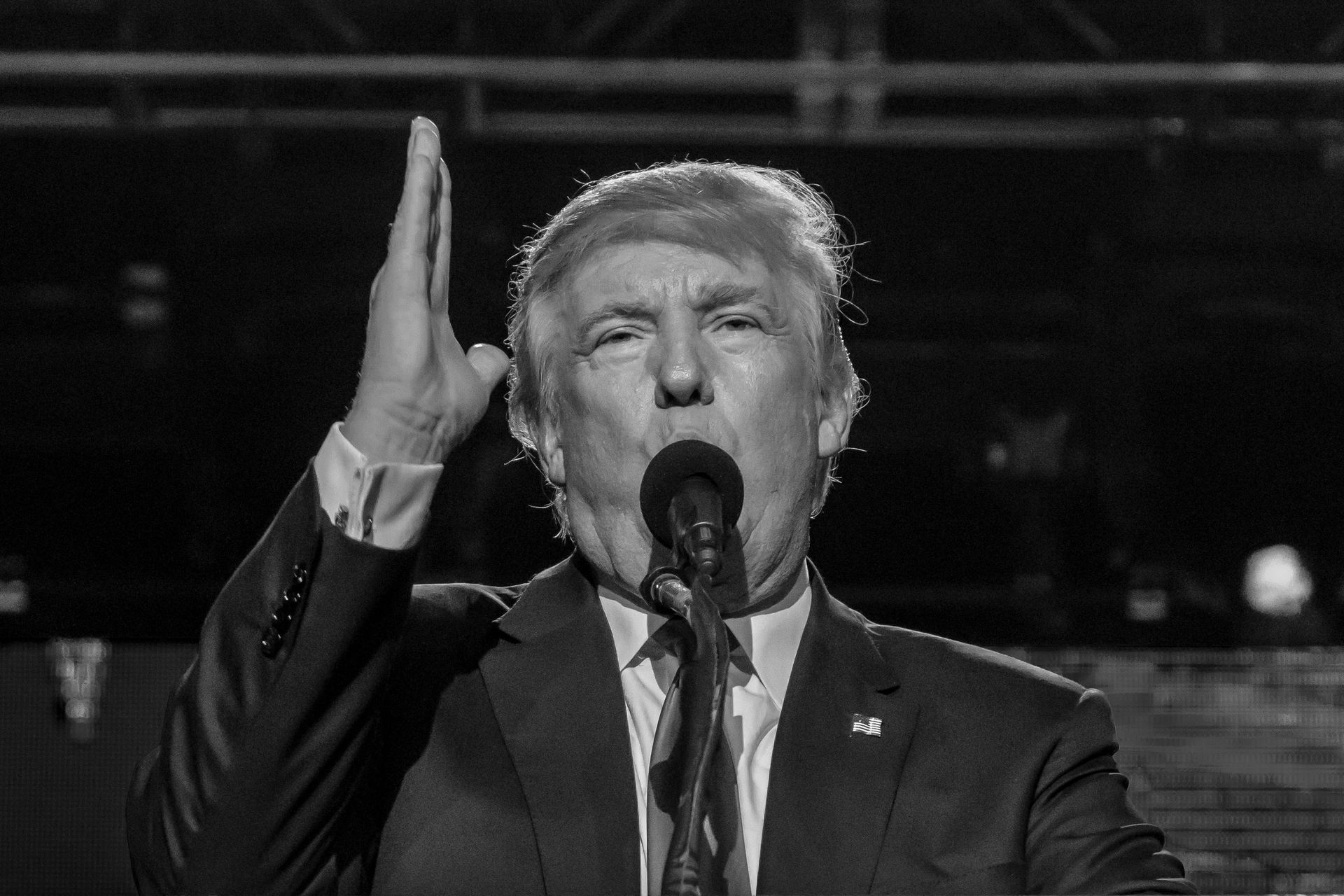 Donald Trump speaking into a microphone, raising his right hand. Black and white photo.
