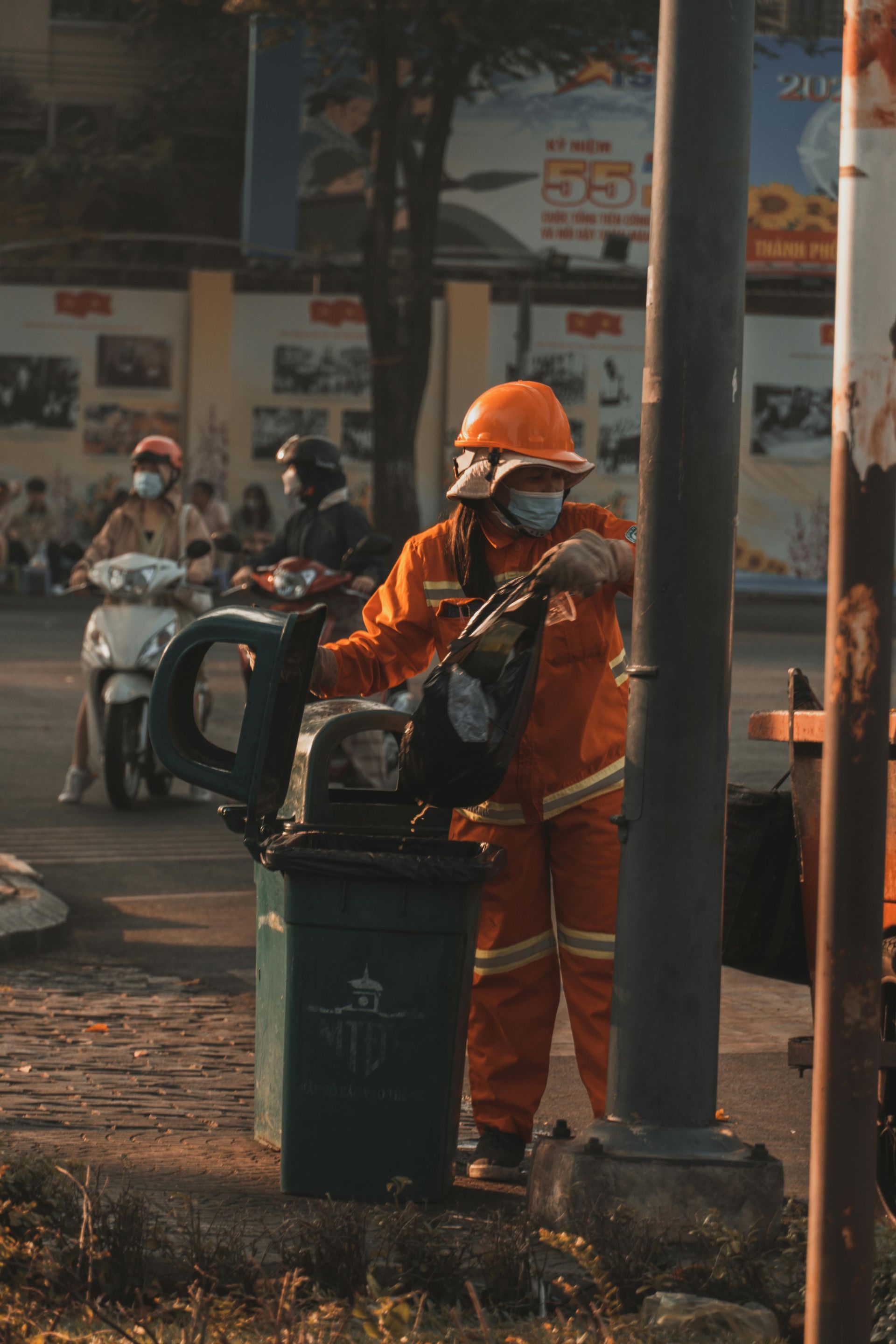 A man in orange overalls and a hard hat is standing next to a trash can.