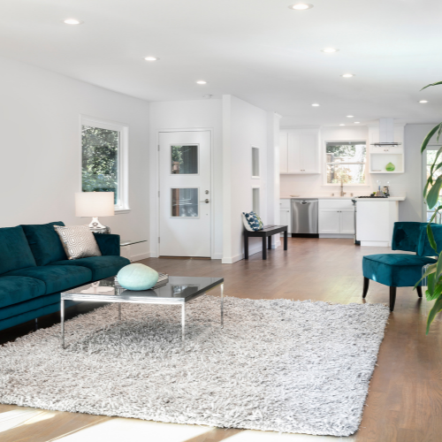 Spacious, modern living room with teal sofa, rug, and open kitchen. White walls and wood flooring.