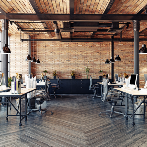 Open-plan office with brick wall, wooden beams, desks, chairs, and hanging lights.