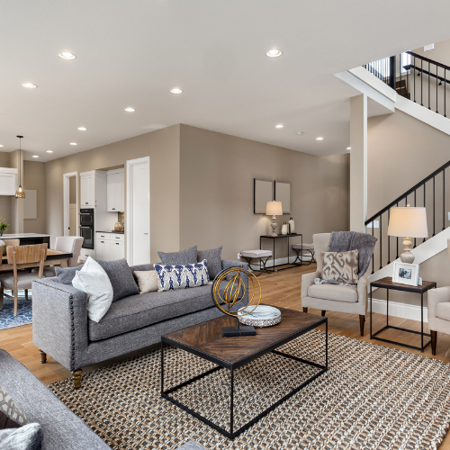 Living room with gray sofa, neutral walls, wood floor, open to kitchen and staircase.