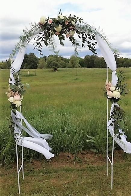 Floral wedding arch with white drapes standing in a grassy field under a cloudy sky