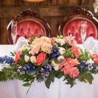Floral wedding table centerpiece with pink, peach, and blue flowers in front of red velvet chairs