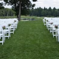 Outdoor wedding aisle with white chairs lining a grassy path beside a pond and trees