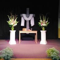 Church altar with cross draped in white cloth, floral arrangements, and a small table on a stage