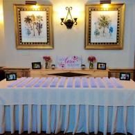 White-covered table set for an event beneath framed floral art and a wall sconce in a softly lit room