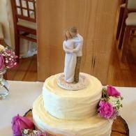 White tiered cake topped with a bride and groom figurine, decorated with pink flowers on a wooden table